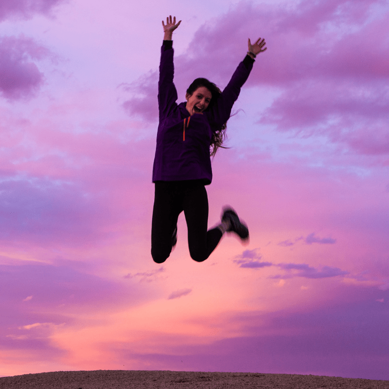 Woman leaping into the air with her hands thrown up into the sky infront of a purple and pink sunset