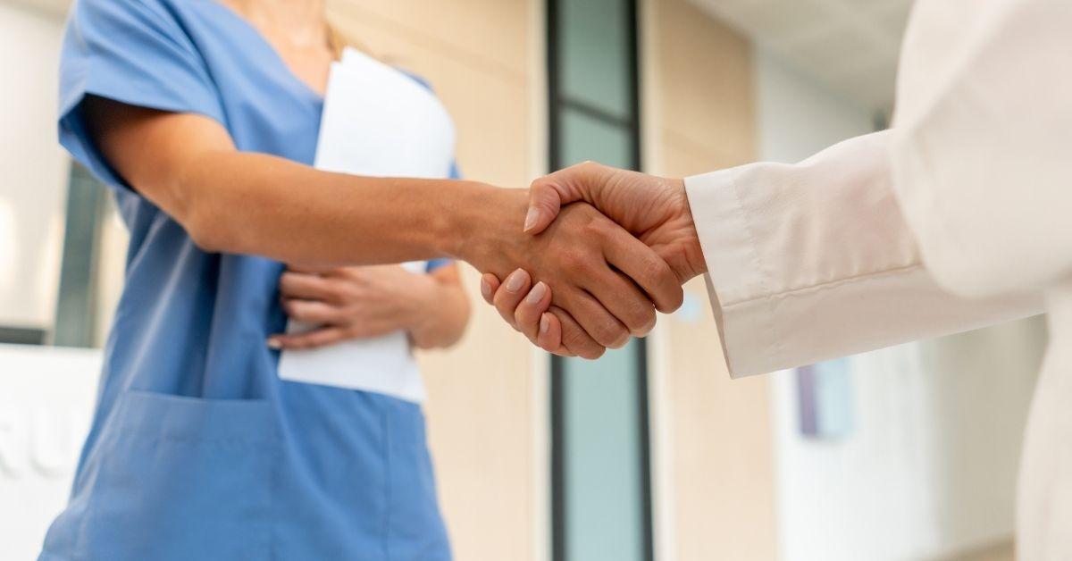 Woman wearing blue scrubs, ho;ding a file folder in left arm, while shaking hands with someone wearing a white jacket, with her right hand